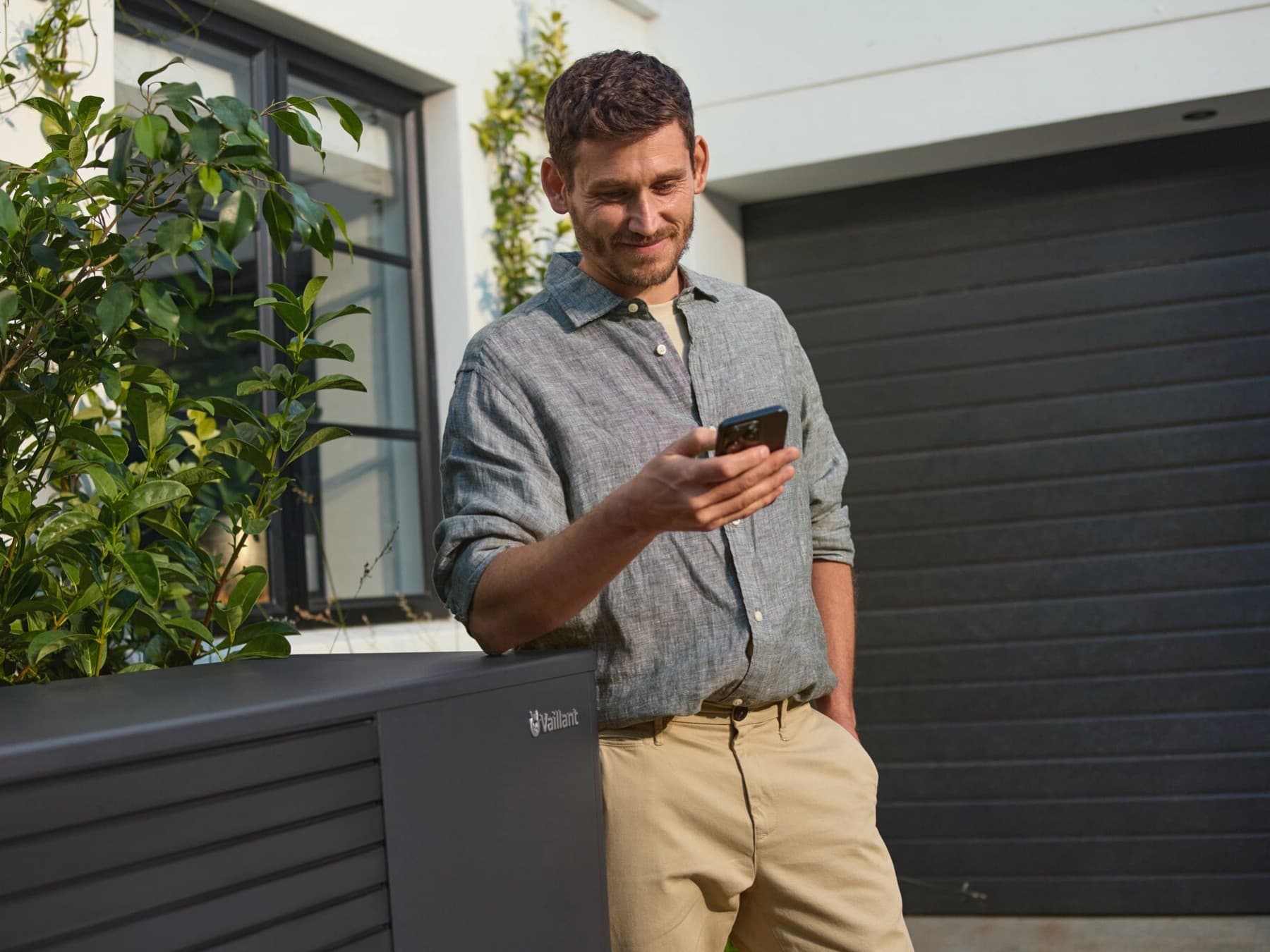 Mann mit Smartphone in der Hand lehnt an Vaillant Wärmepumpe vor einem Haus